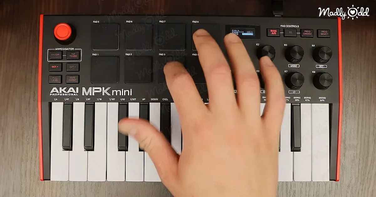 A hand presses a key on an Akai MPK Mini MIDI keyboard with pads and knobs visible.