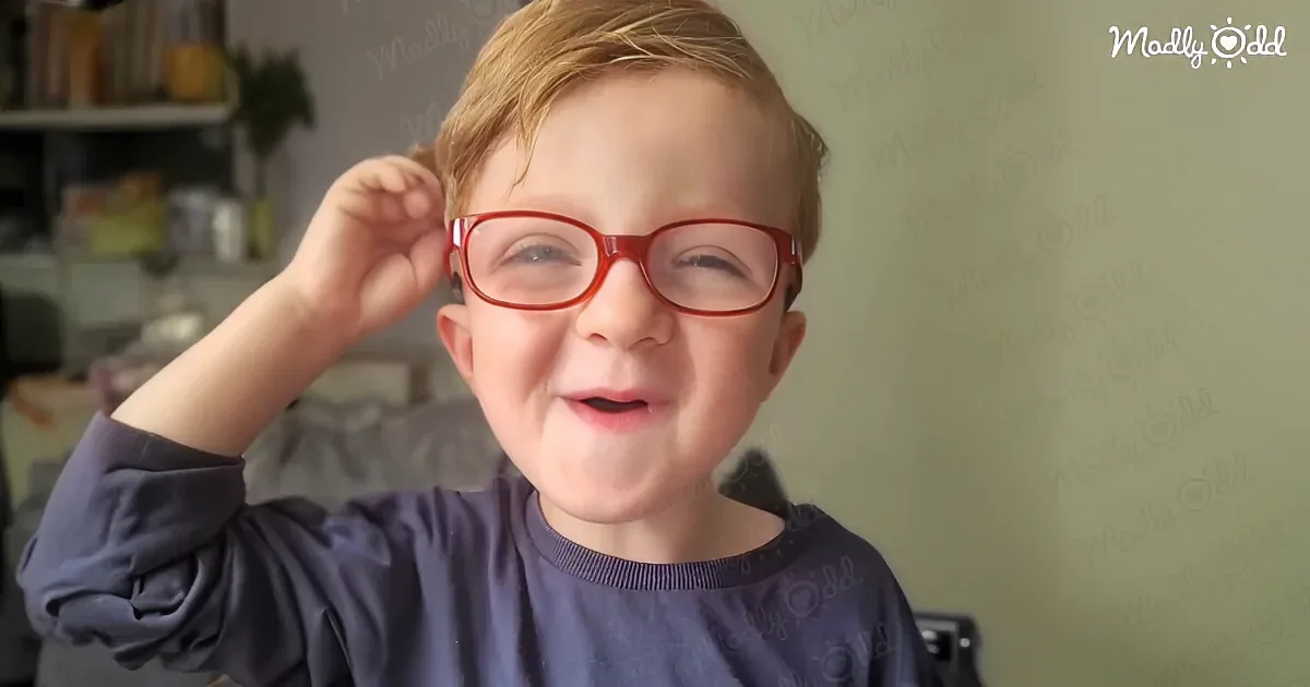 A young boy with red glasses smiles, adjusting his glasses with one hand.