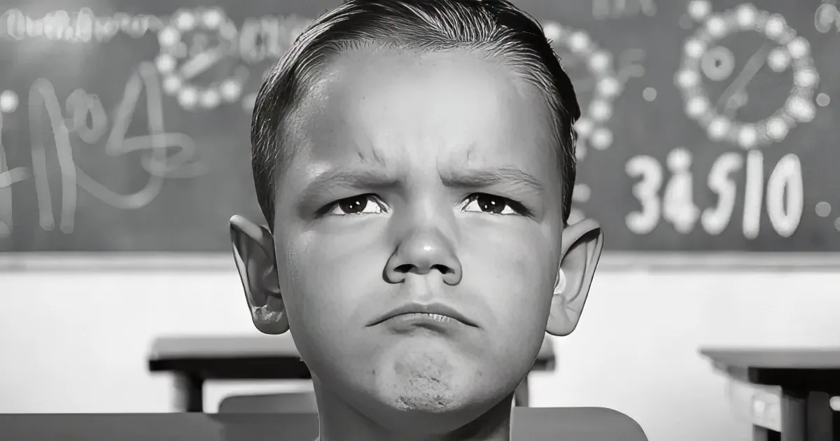A young boy with a furrowed brow and a concerned expression sits in front of a chalkboard.