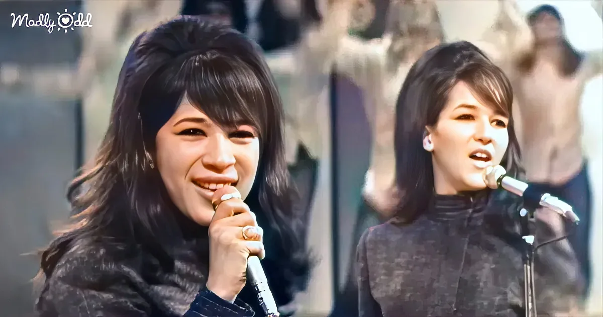 A woman with long dark hair sings into a microphone, smiling, with another woman in the background.