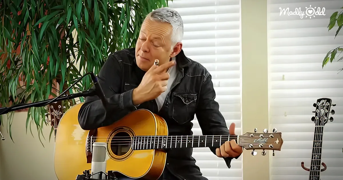 A man in a black jacket plays an acoustic guitar, holding a cigarette, with a microphone and plants in the background.