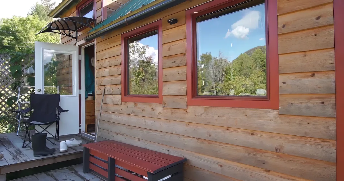 A cozy wooden cabin with a solar panel, surrounded by lush greenery and vibrant flowers, bathed in sunlight.