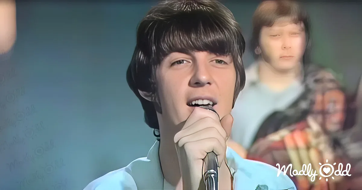 A young man with dark hair and bangs sings into a microphone, wearing a light blue shirt.