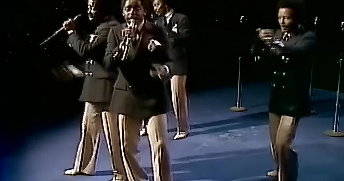 A man in a suit sings into a microphone, surrounded by confetti against a dark background.