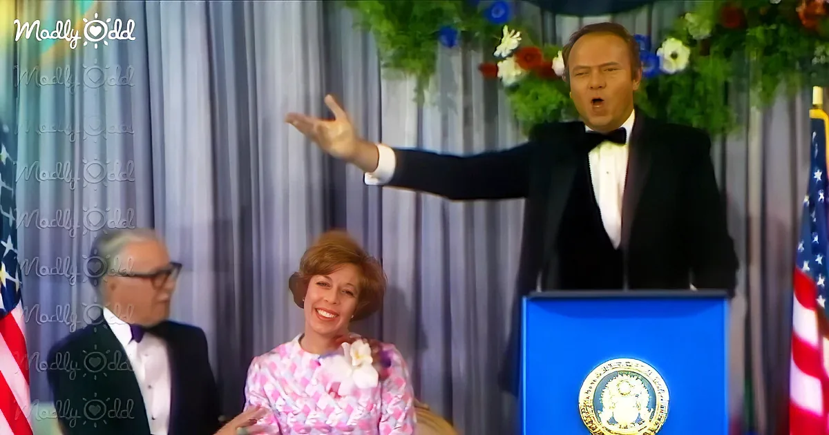 A man in a tuxedo gestures dramatically at a podium, with a woman in a floral dress and another man in a suit in the...