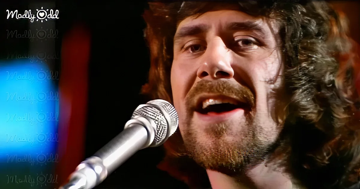A man with curly hair and a beard sings into a microphone, his eyes focused, against a blurred background.