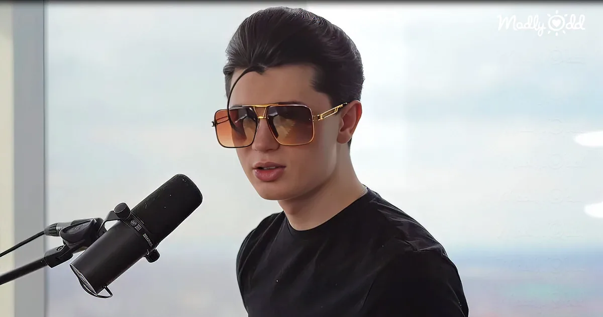 A person wearing sunglasses and a black shirt stands in front of a microphone, with a blurred background.