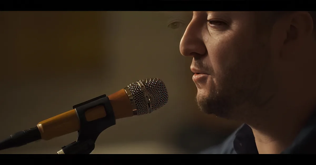 A man with a beard sings into a microphone, his eyes closed, in a warmly lit setting.