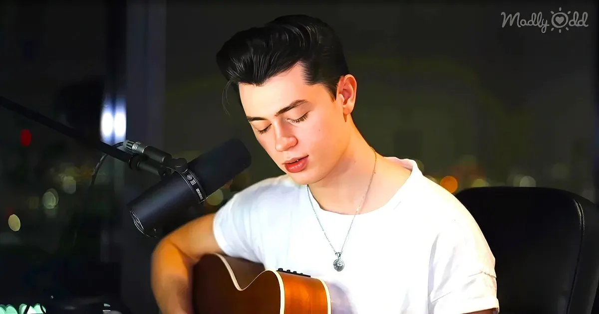 A person in a white shirt plays an acoustic guitar, singing into a microphone, with a blurred background.
