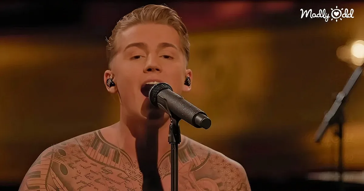 A person with tattoos sings into a microphone, wearing earpieces, against a blurred background.