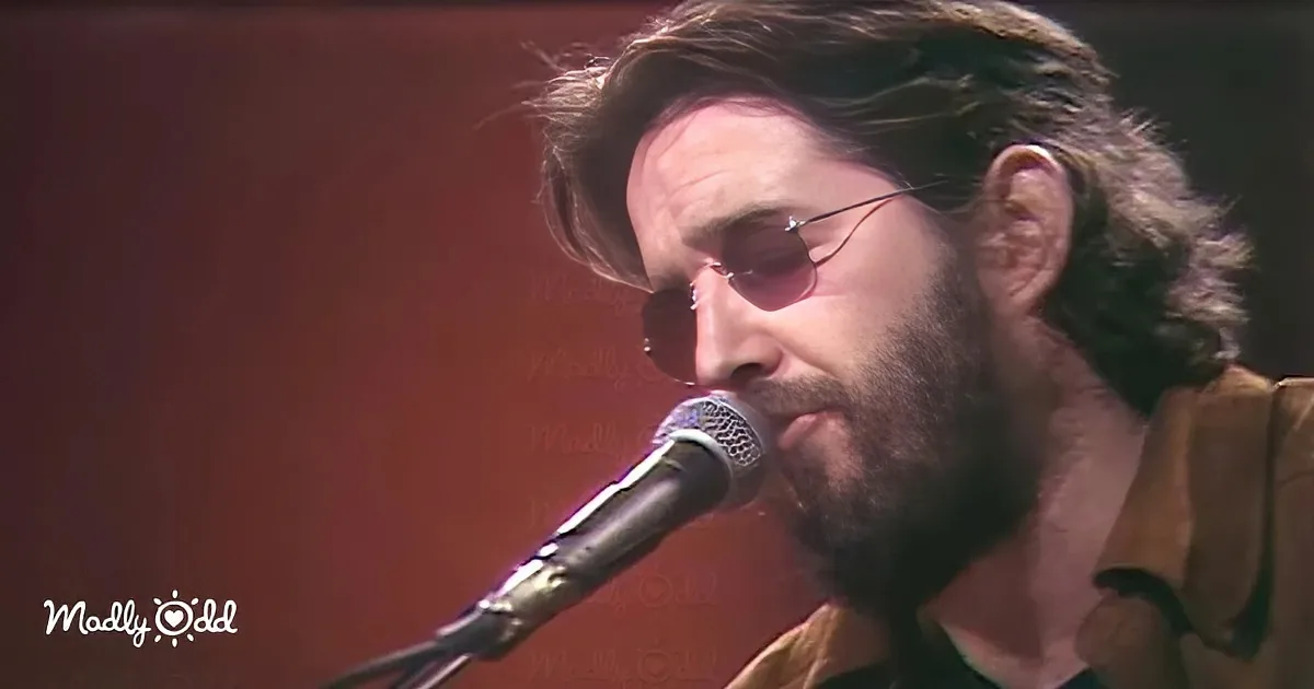 A man with sunglasses and a beard sings into a microphone, wearing a brown shirt.