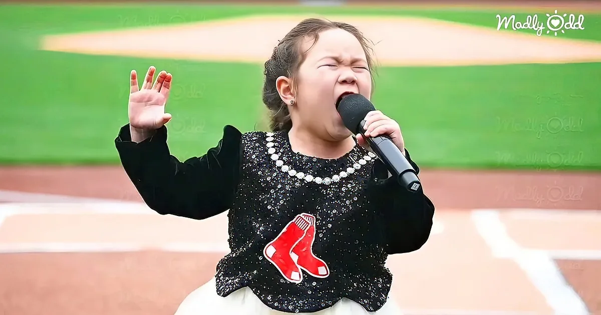 A young girl in a black sequined top with a red logo sings into a microphone on a baseball field.