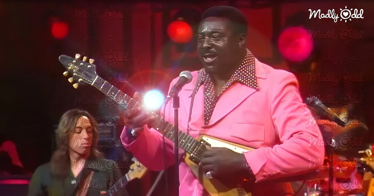 A man in a pink suit plays guitar and sings on stage, illuminated by red lights.