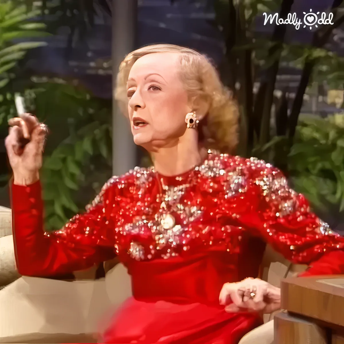 Bette Davis with cigarette in red dress sitting on Carson set.