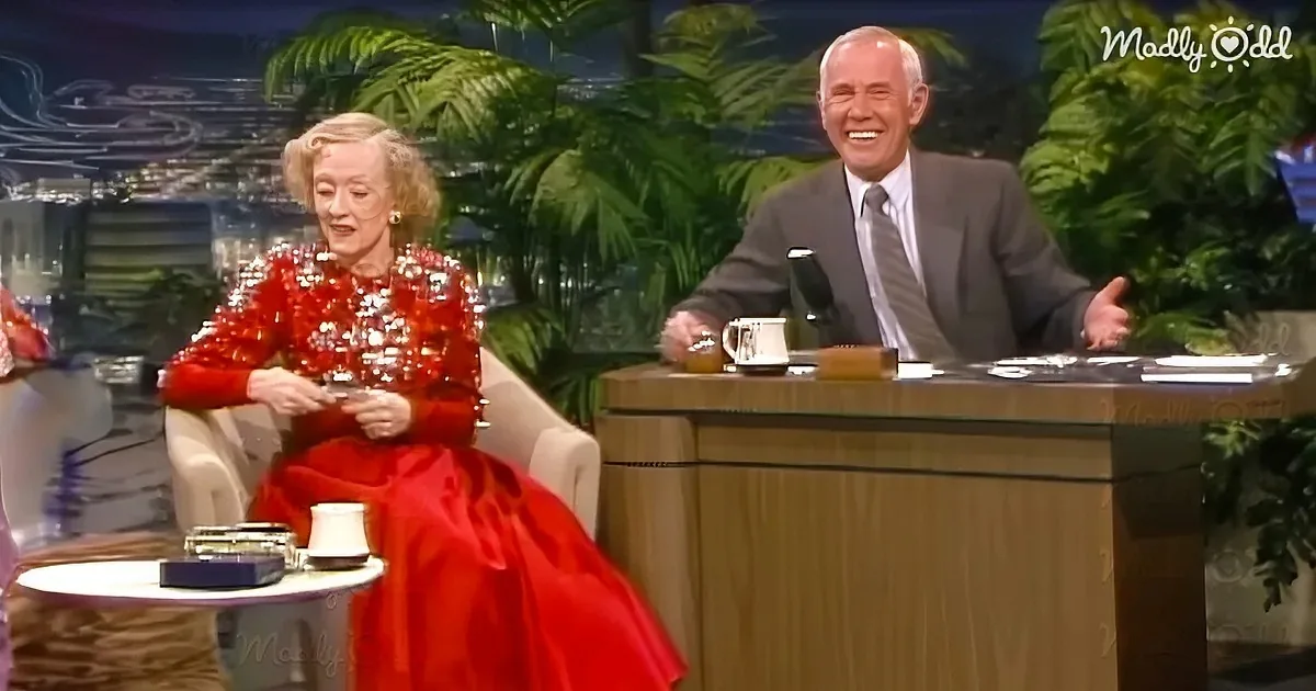 Woman in red dress and man in suit on a talk show set with tropical plants in the background.