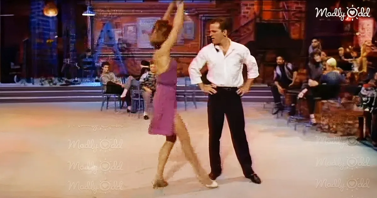 A man in a white shirt and black pants stands with hands on hips, watching a woman in a purple dress perform a dance move.
