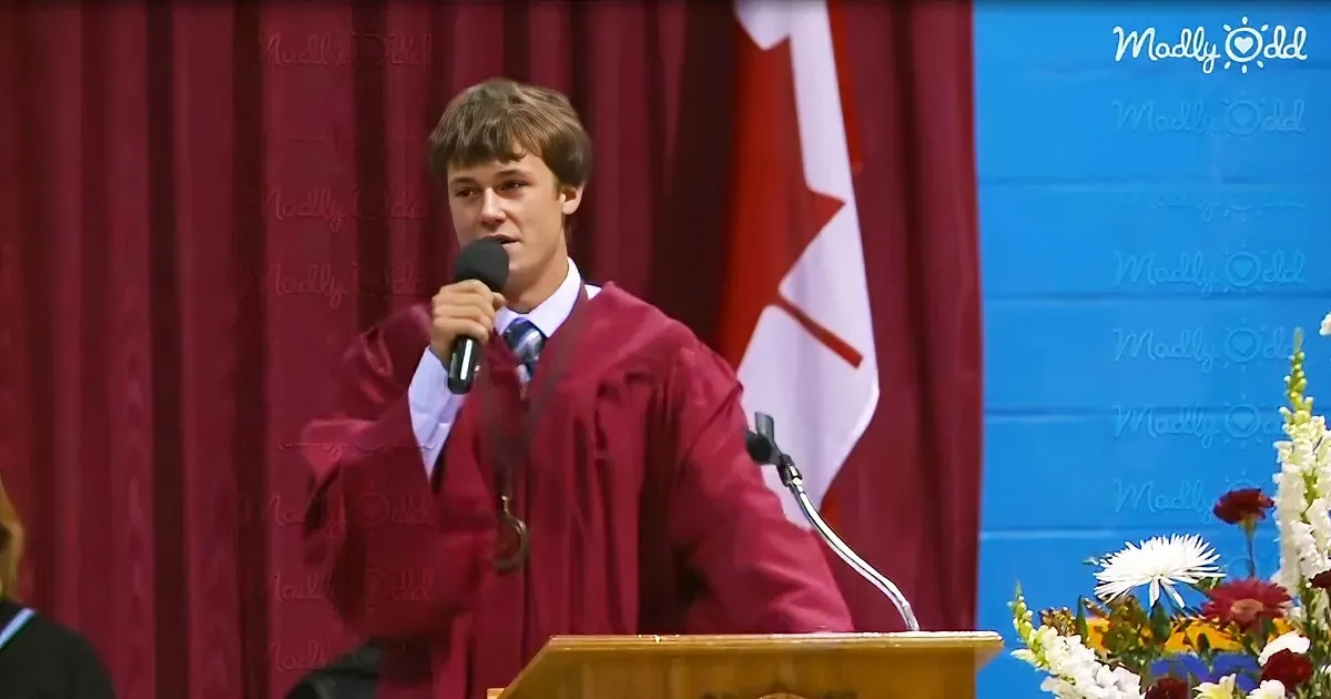A young man in a maroon graduation gown speaks into a microphone at a podium.