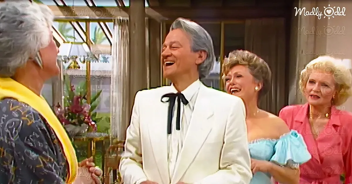A man in a white suit with a black bow tie laughs heartily, surrounded by smiling women in elegant attire.