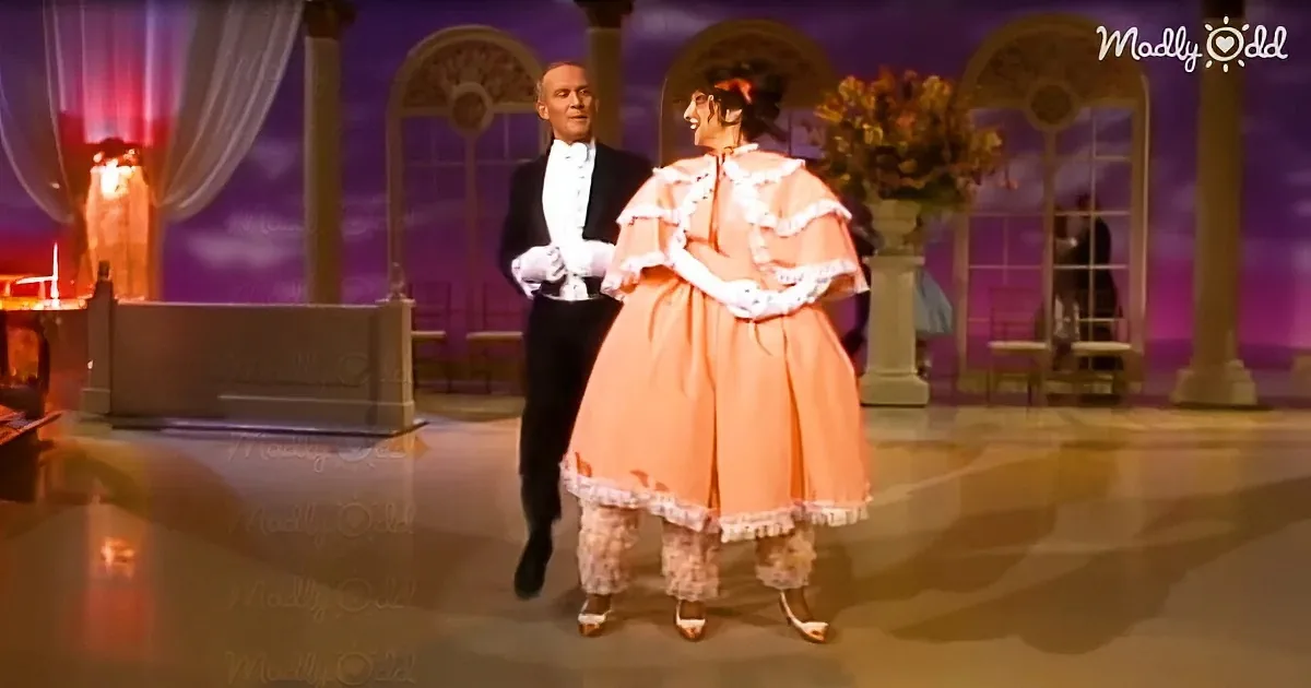 A man in a tuxedo and a woman in a peach-colored dress are dancing on a stage with a purple backdrop and decorative elements.