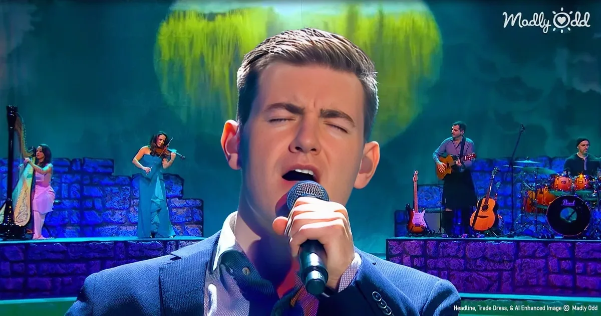 A man sings passionately into a microphone on stage, surrounded by musicians and a scenic backdrop.