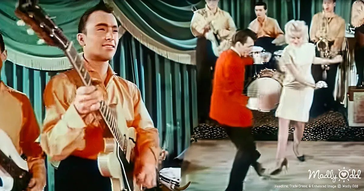 A man in an orange shirt plays guitar while a band and dancers perform in the background.