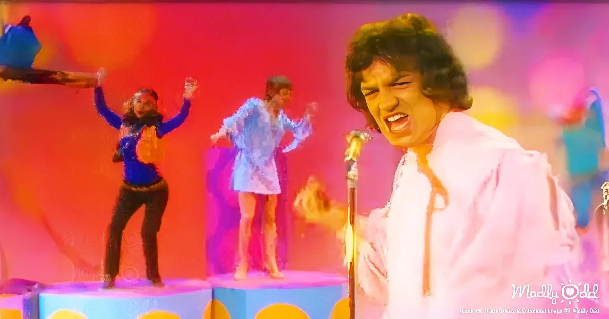 A man in a pink shirt sings passionately on stage with colorful, vibrant background and dancers.