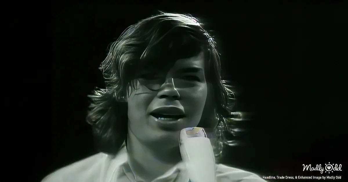 A person with long hair and glasses is singing into a microphone, illuminated by a spotlight.