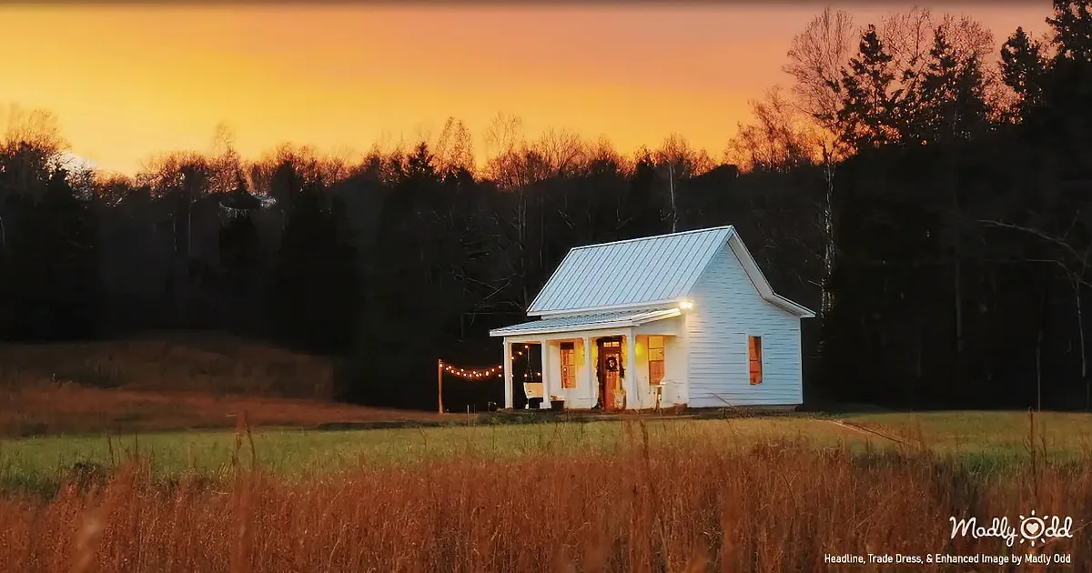 A cozy white house with string lights glows warmly against a vibrant orange sunset backdrop.