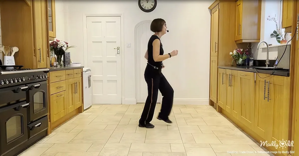 A woman in black attire dances in a kitchen with wooden cabinets and a black oven.