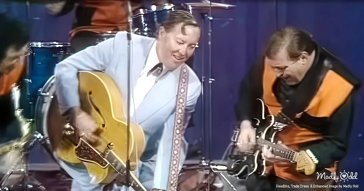 A man in a white suit plays a yellow guitar, smiling, with another person in an orange jacket playing a black guitar nearby.