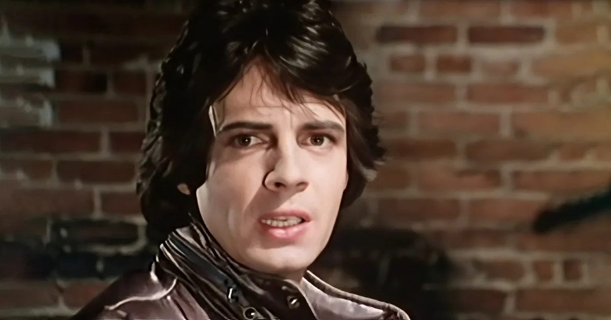 A person with dark, wavy hair and a leather jacket stands against a brick wall, looking to the side.
