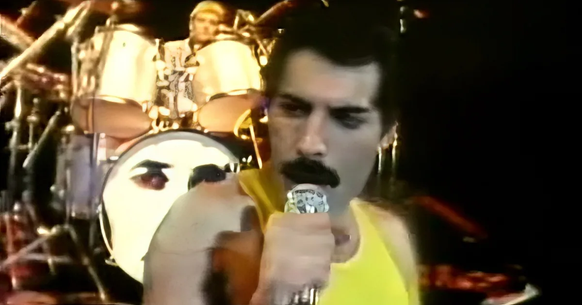 Freddie Mercury sings passionately on stage, holding a microphone, with a drummer in the background.