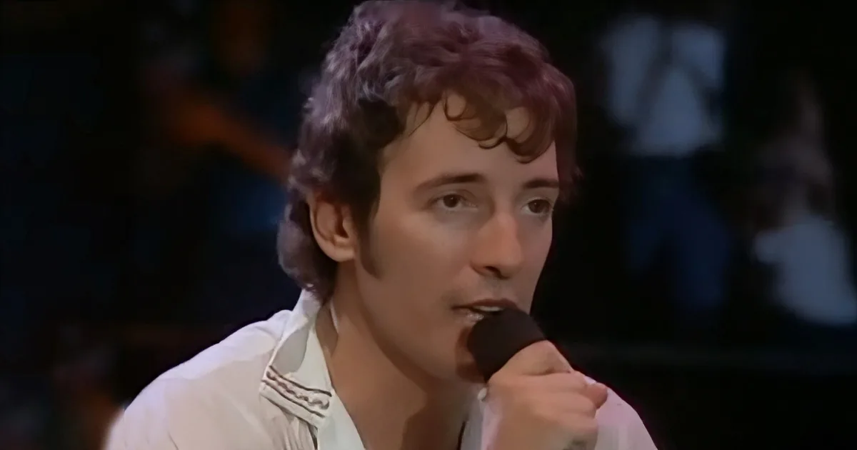 A man with curly hair in a white shirt sings into a microphone.