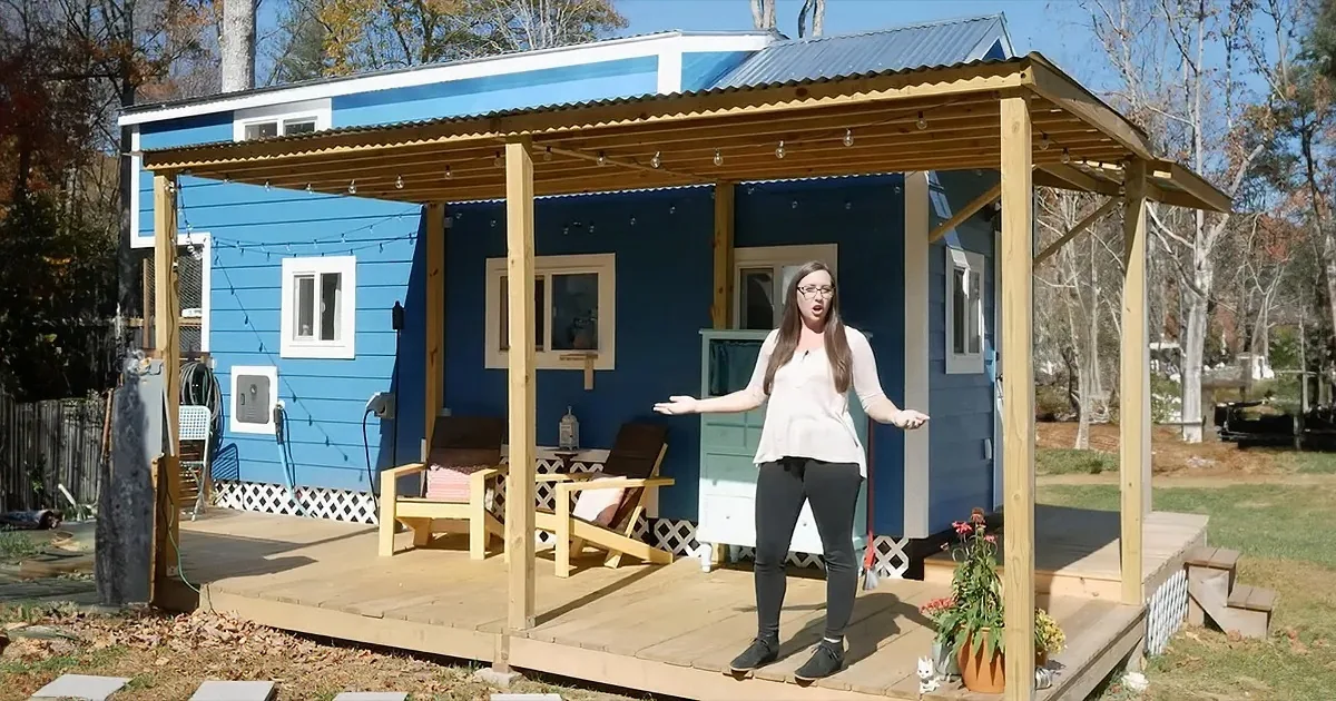 A woman stands on a wooden deck in front of a blue tiny house with a covered porch and string lights.