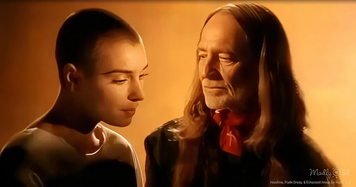 A man with long hair and a red tie stands beside another man with a shaved head, both in a dimly lit setting.