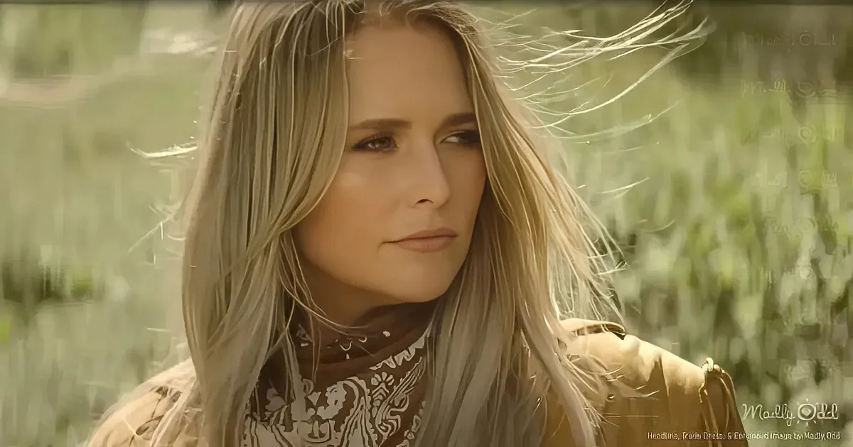 A woman with long blonde hair, wearing a patterned top and a beige jacket, stands in a grassy field with her hair blowing...
