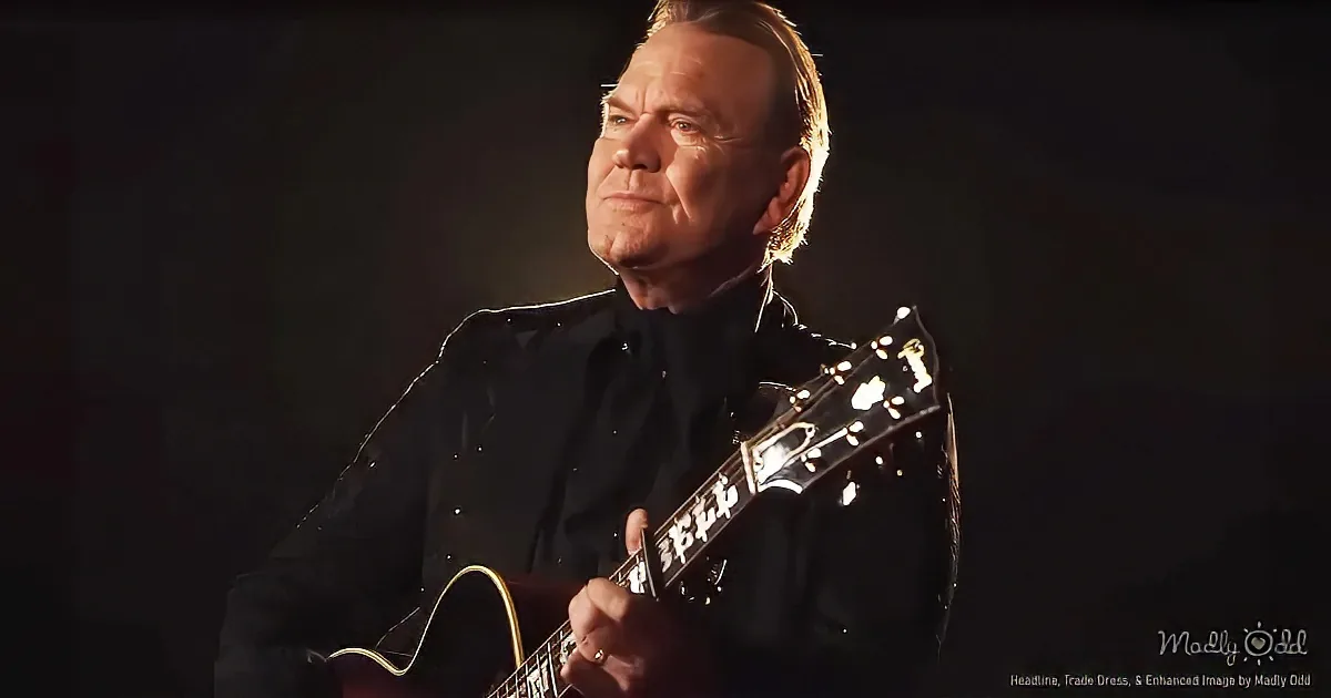 A man in a black suit plays a guitar under focused lighting, creating a dramatic effect.