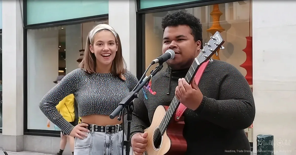 A man plays guitar while a woman smiles nearby, both in front of a store window.