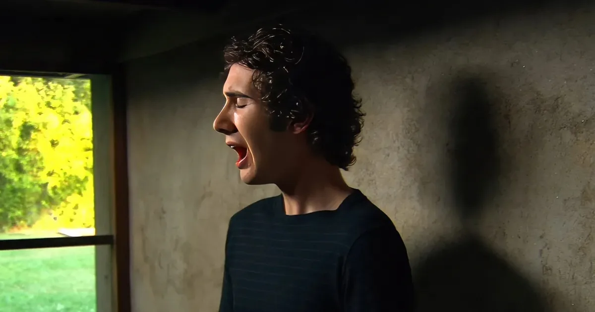A person with curly hair stands indoors, mouth open, looking out a window with greenery visible outside.