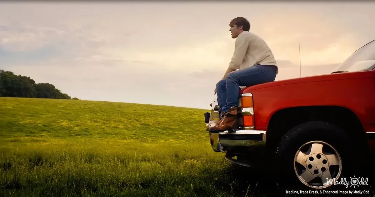A man sits on a red truck in a grassy field, gazing into the distance under a cloudy sky.