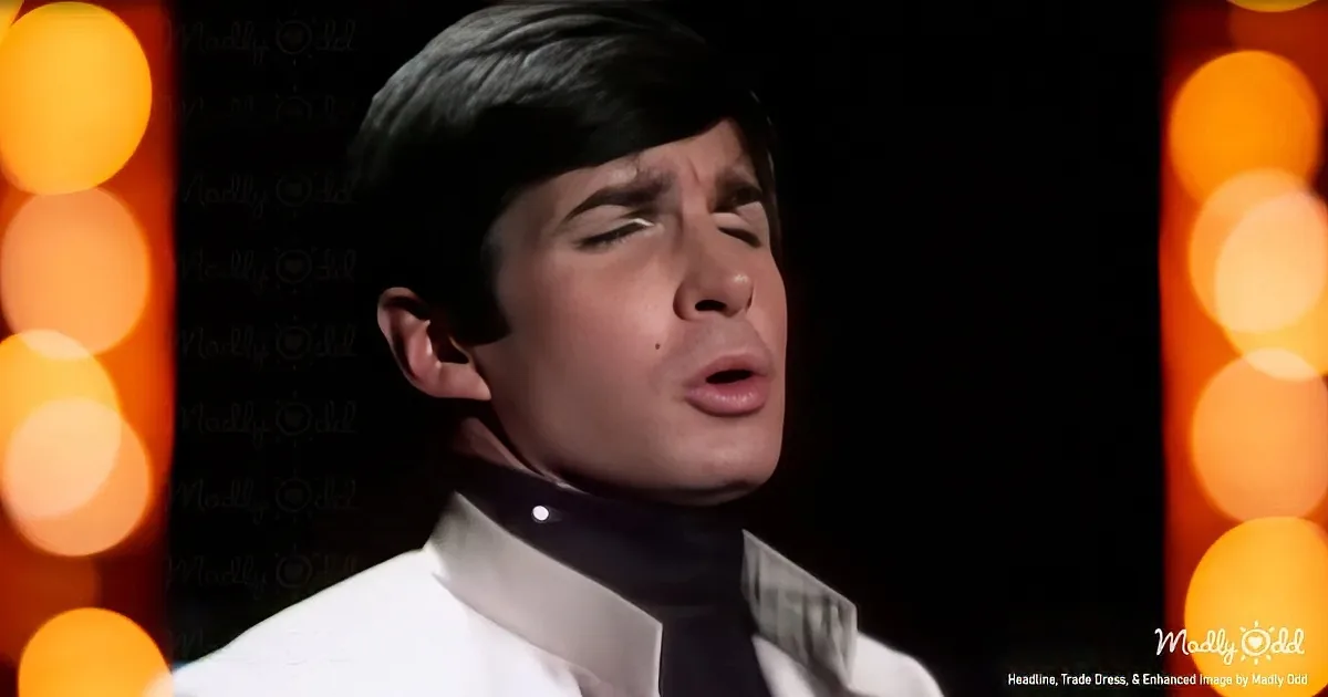 A man in a white shirt and black tie sings with eyes closed, surrounded by blurred orange lights.