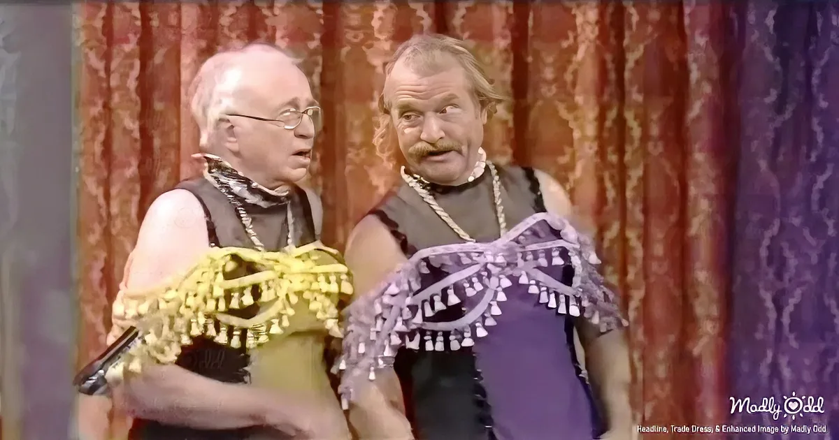 Two men in colorful, fringed costumes stand in front of a patterned curtain, one holding a microphone.