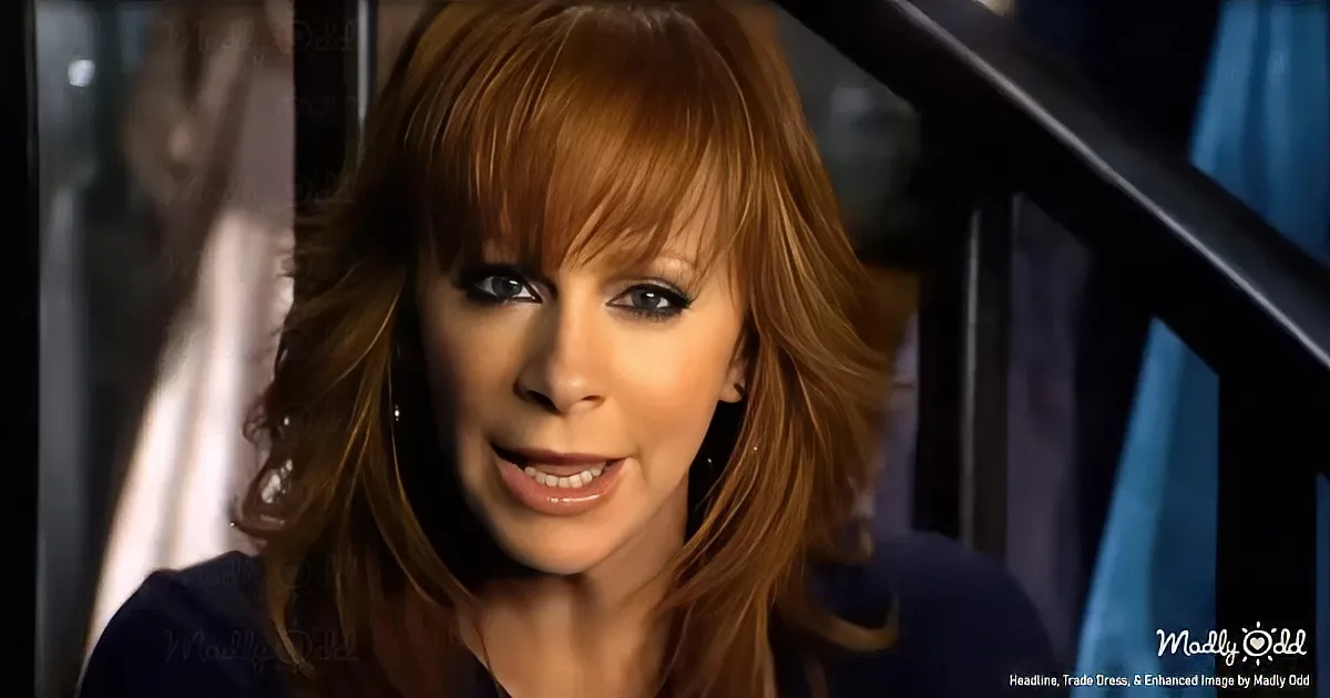 A woman with long, wavy brown hair and bangs, wearing a dark top, smiles slightly in a dimly lit setting.