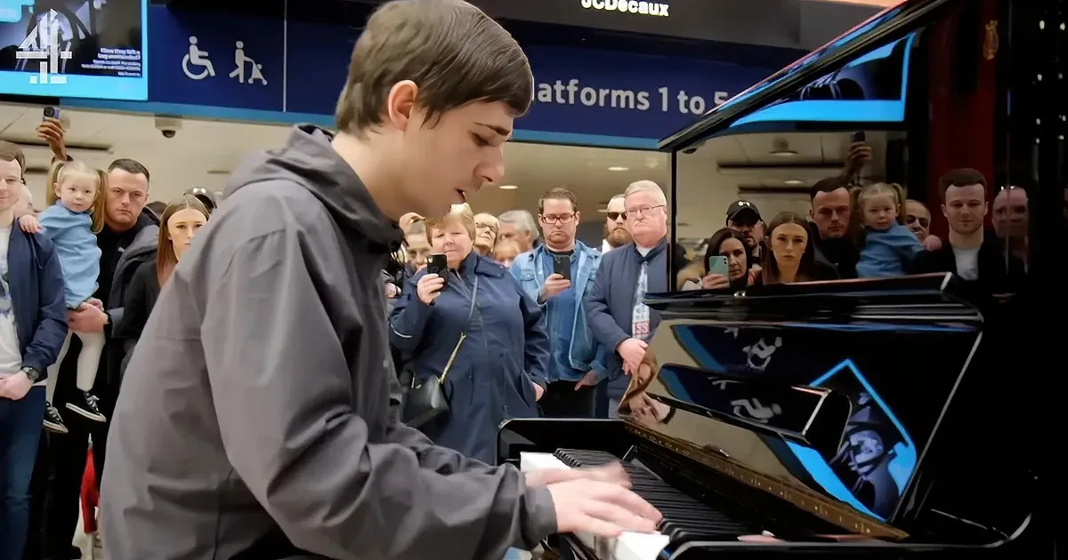 A young man in a gray jacket plays a grand piano in a crowded station, surrounded by onlookers.