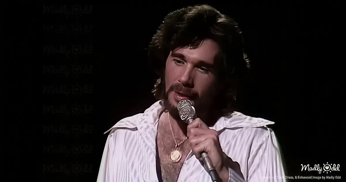 A man with long hair and a mustache holds a microphone, wearing a white shirt with a gold necklace, against a dark...