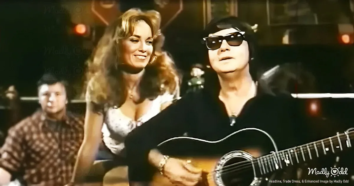 A man in sunglasses plays guitar, smiling woman in white dress behind him, other people in background.