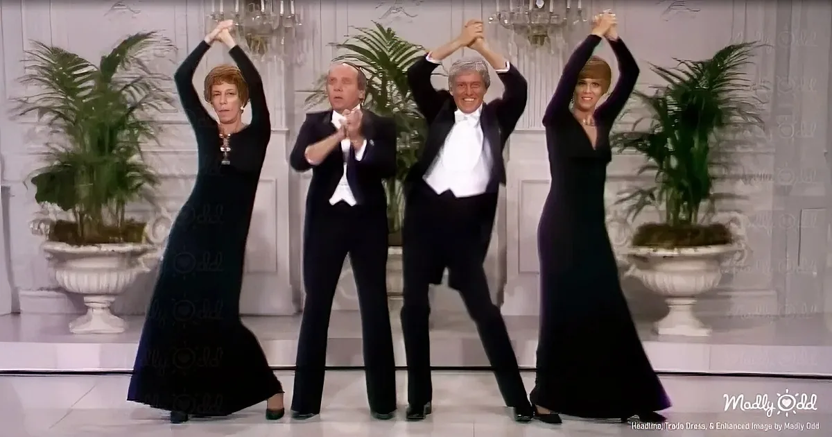 Four people in formal attire dance together on a stage with chandeliers and potted plants in the background.