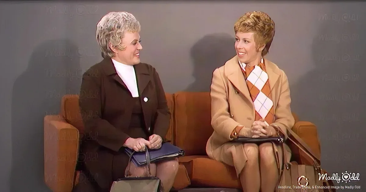 Two women in vintage attire sit on a couch, engaged in conversation, with one holding a handbag.