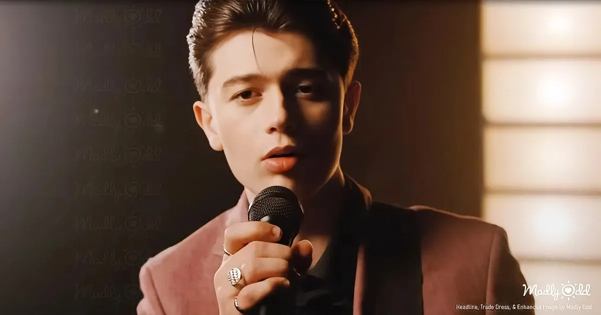 A person in a pink suit holds a microphone, wearing a ring, with a dark background and soft lighting.