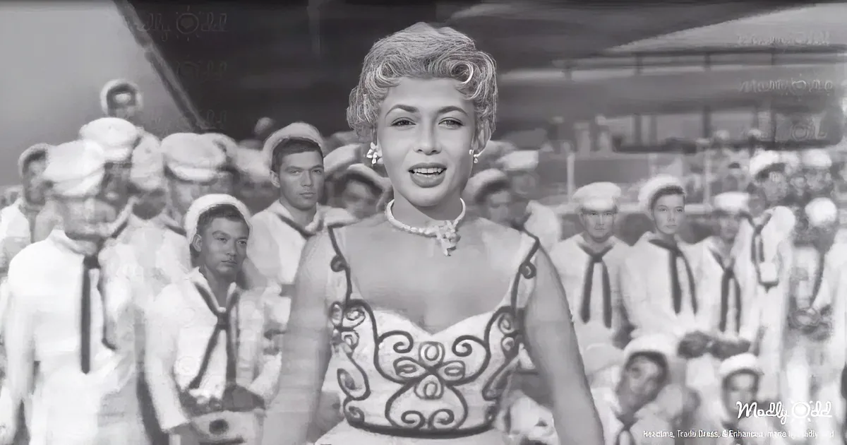 A woman in a white dress with a floral pattern sings in front of a group of sailors in white uniforms.